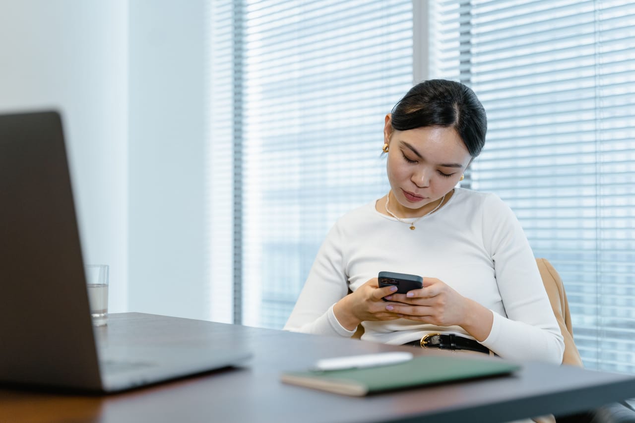 A woman reading her phone at an office desk, with a laptop in the foreground