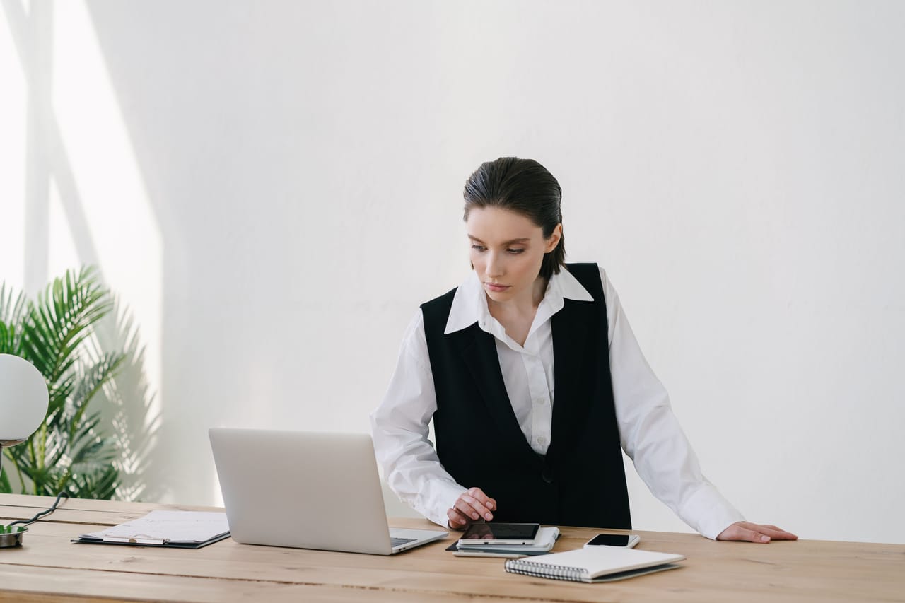 A woman standing at a wooden desk, using a tablet beside her laptop
