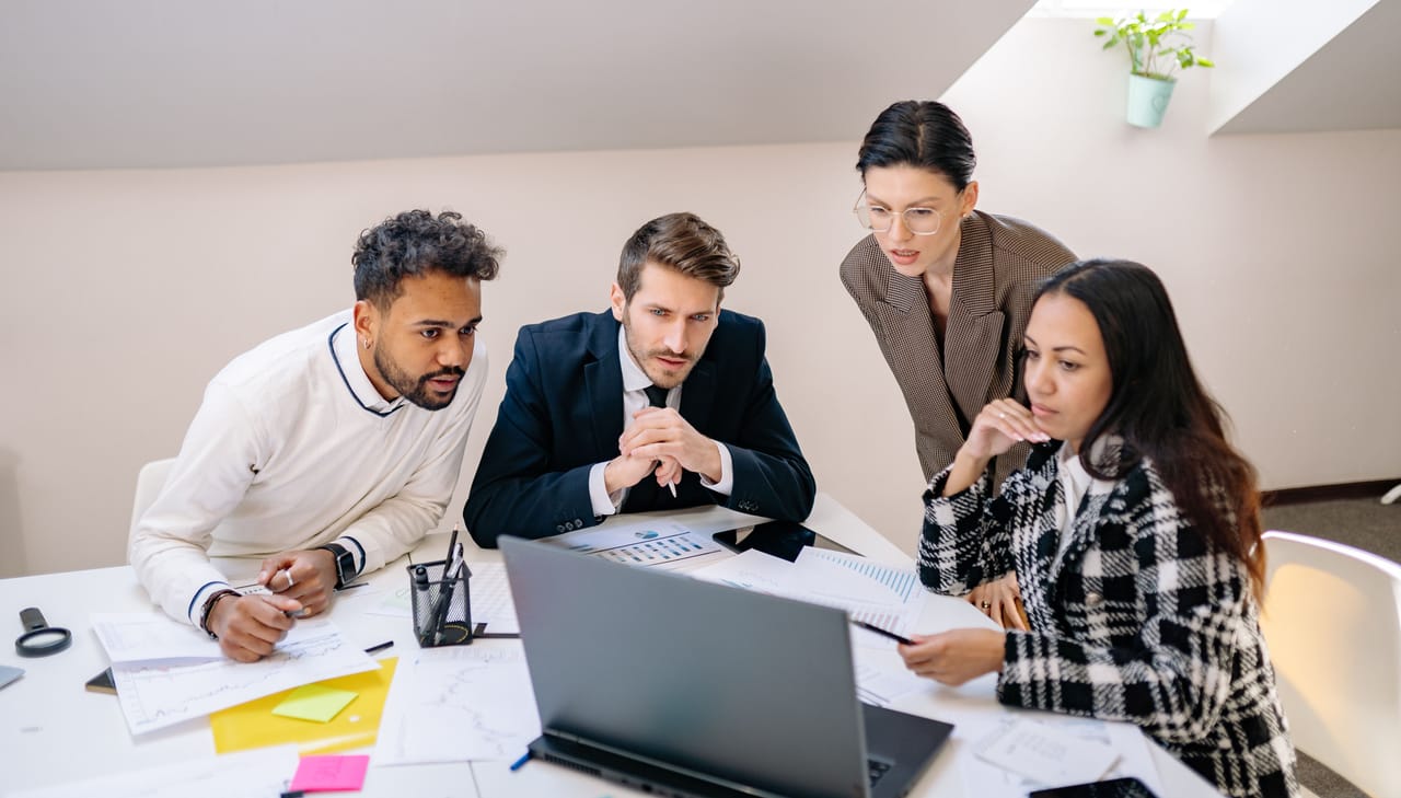 Four colleagues gathered around a laptop at a meeting table covered with charts