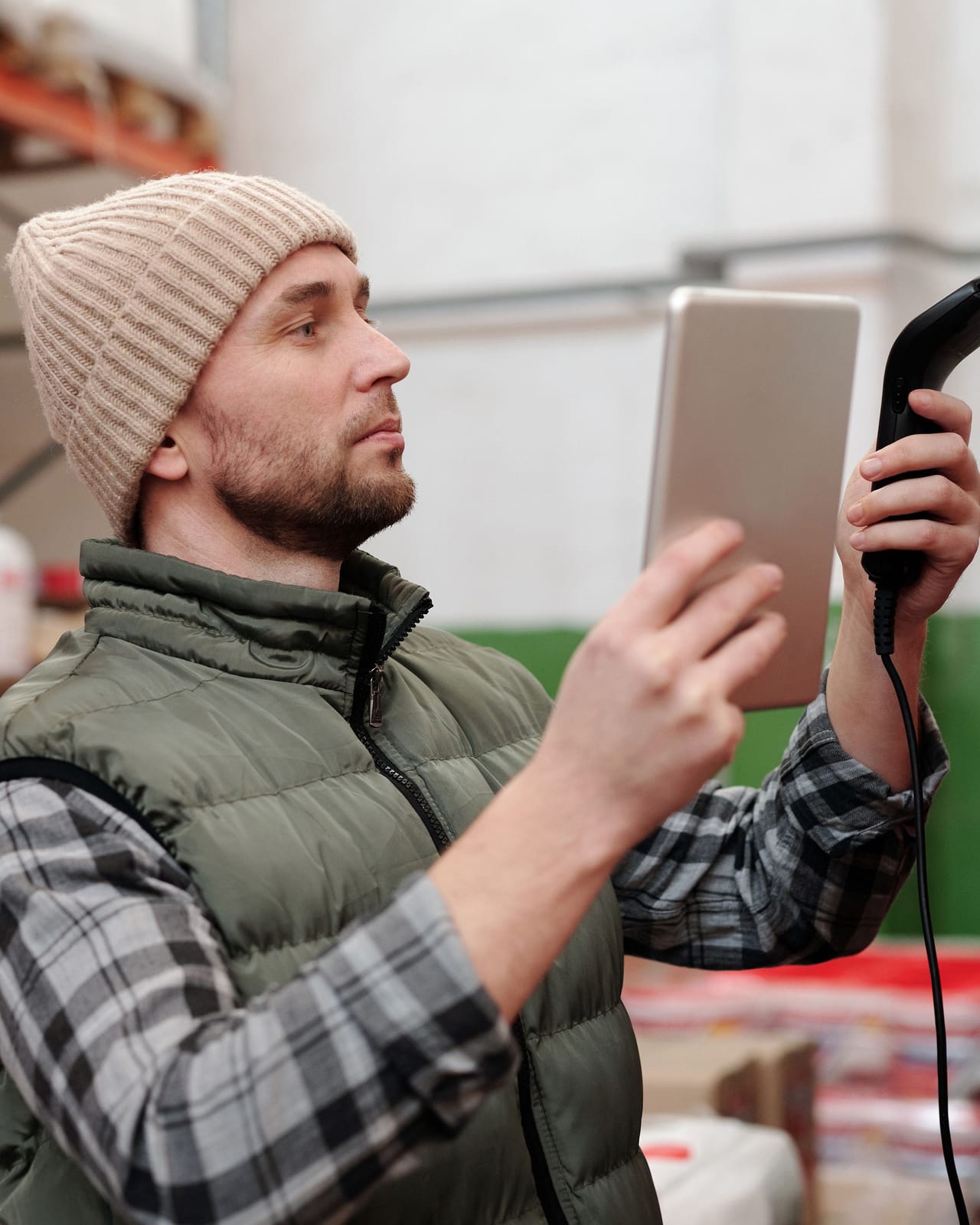 A warehouse worker in a knit hat checking stock with a handheld scanner and a tablet